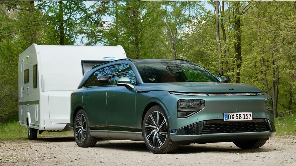 Bild av en grön elbil som drar en vit husvagn, parkerad på en skogsväg med blommor i förgrunden.