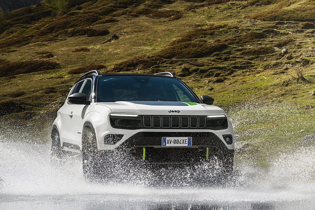 Vit Jeep kör genom vatten med en gräsbevuxen kulle i bakgrunden.