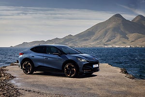 Blå Cupra parkerad vid en klippa med havet och bergen i bakgrunden.