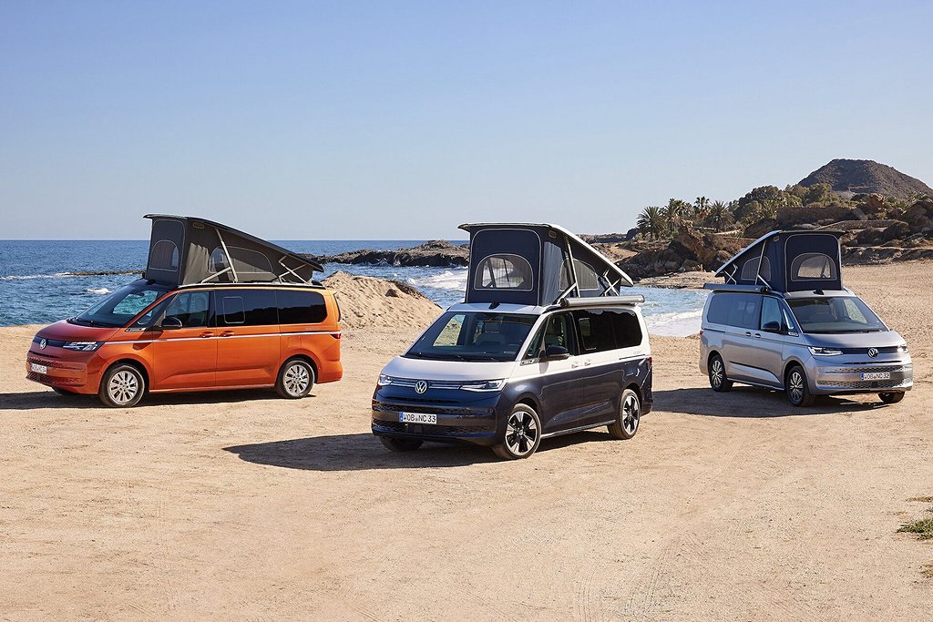 Tre Volkswagen camping med uppfällda tak parkerade på en sandstrand med havet och klippig kustlinje i bakgrunden.